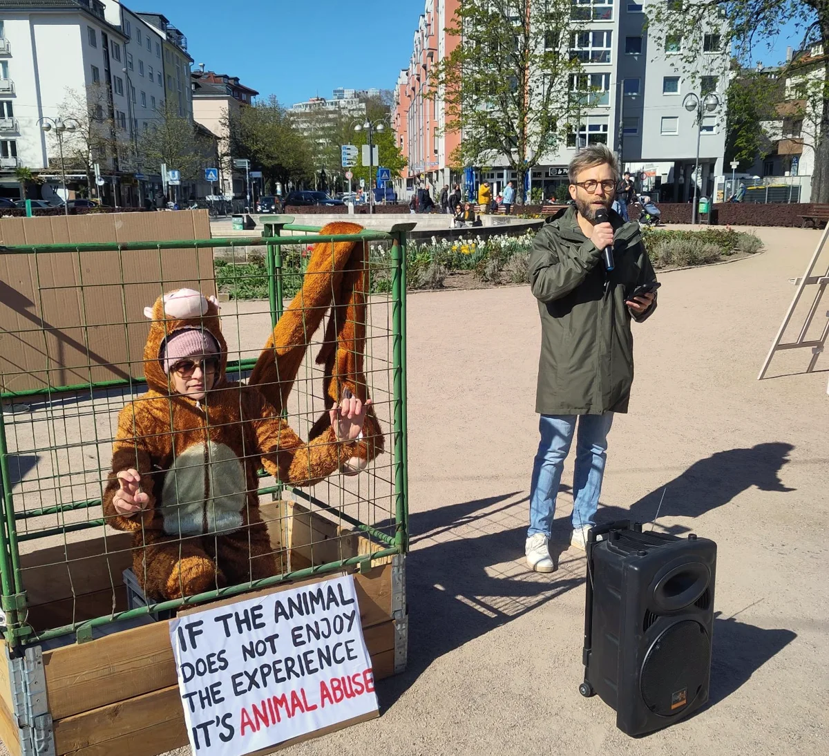 Sven Ganter spricht mit Mikrofon bei einer Mahnwache vor dem Frankfurter Zoo, daneben eine Aktivistin im Affenkostüm in einem Käfig